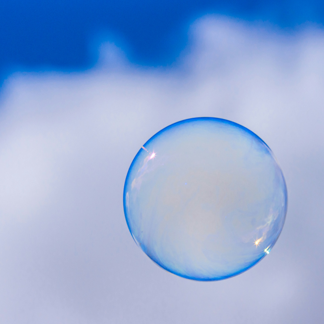 An abstract photo of an iridescent blue bubble floating in front of a white cloud in a blue sky.